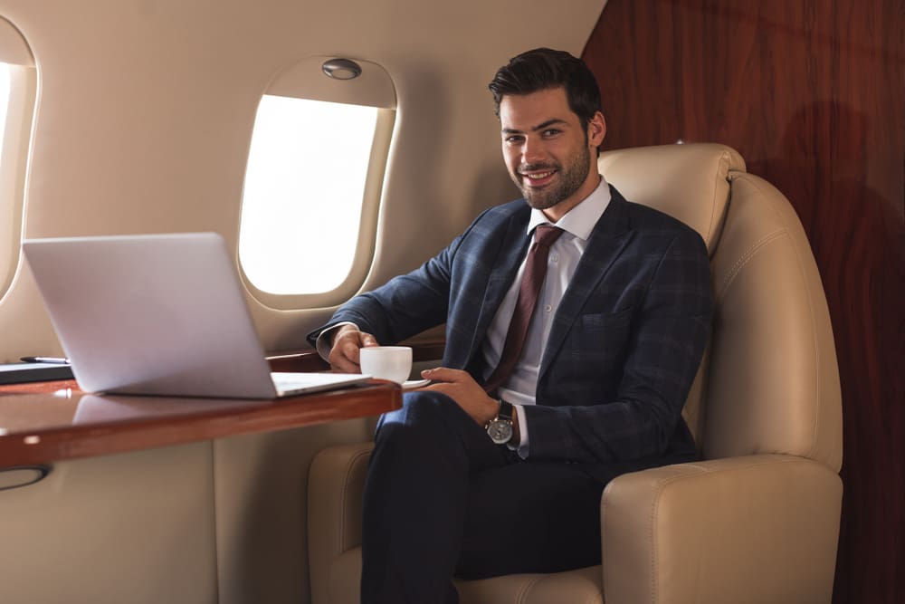 Smiling,Businessman,Holding,Cup,Of,Coffee,In,Plane,With,Laptop